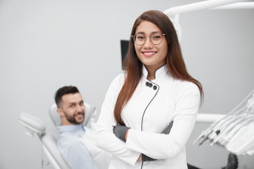 Woman dentist standing with her arms crossed smiling, patient in the background